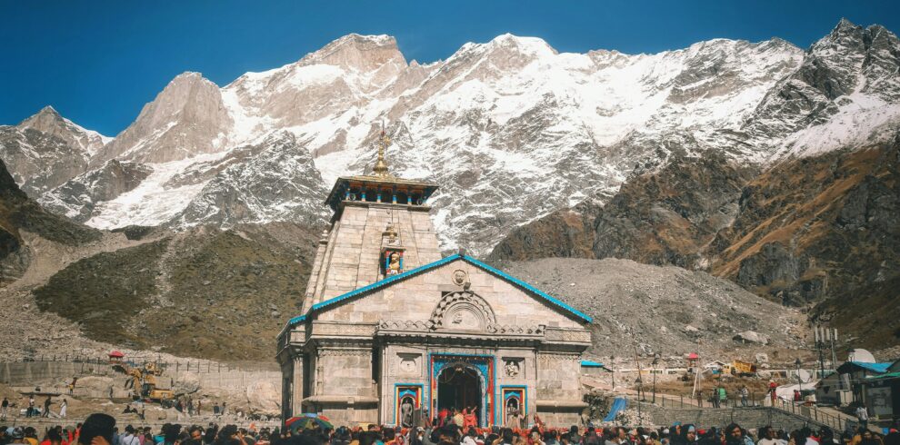 A crowd of pilgrims at Kedarnath Temple with snowcapped Himalayan peaks in the background.